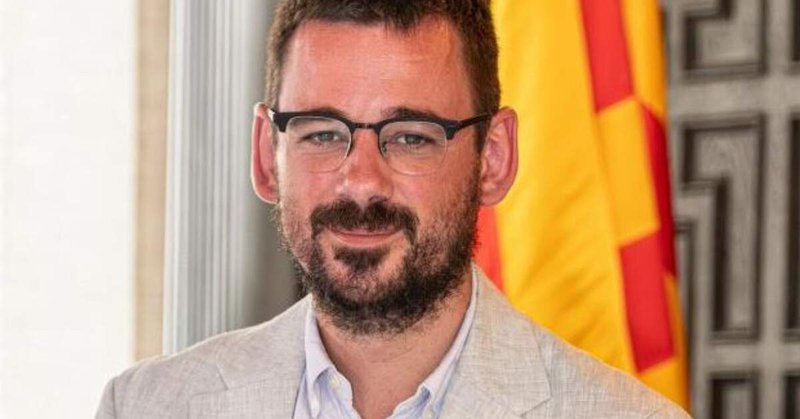 Home amb ulleres i barba vestint americana clara davant d’una bandera groga i vermella