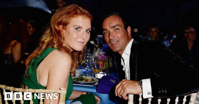 Sarah Ferguson and Manuel Fernandez sit at a table laden with glasses, plates and a blue table cloth. She wears a green dress, he wears a black velvet jacket and a white shirt open at the neck. They sit at wickerwork-style chairs, and are looking over their shoulders at the camera