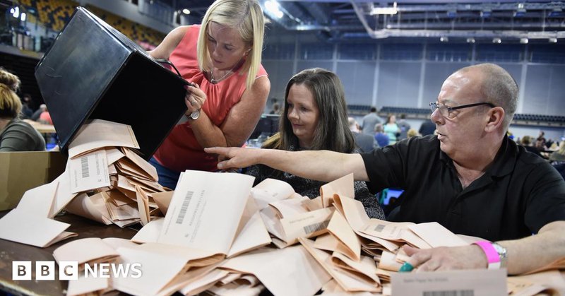 Counting staff check ballot papers in the Scottish Parliament elections at the Emirates Arena, Glasgow, in 2016. A woman on the left is pouring a box of pink papers onto a table, which is covered with ballot papers. A man on the right is pointing at the box and a woman in the middle is looking at the box.