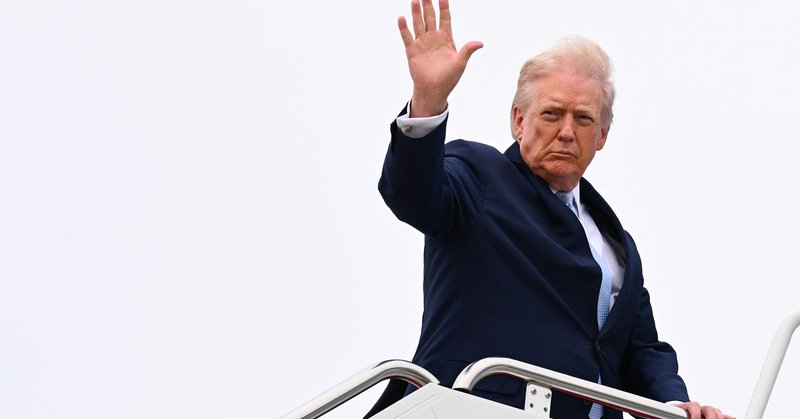 Donald Trump raises his hand while standing on the stairs of Air Force One against an overcast sky.
