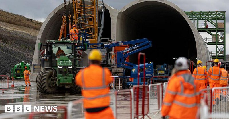 People in hard hats and high vis jackets in soft focus watch as a HS2 tunnel is built using a crane in front of them
