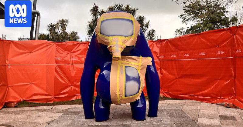 A large blue sculpture with gold tape and grey markings on it in front of an orange fence.