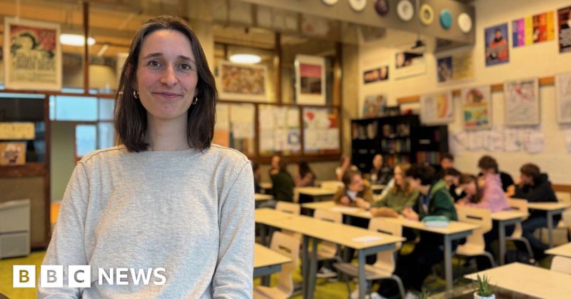 A brunette woman with a bob smiles at the camera in front of a classroom of teenagers. She is smiling and wearing a grey jumper. There are about a dozen children in the class