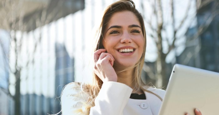 A woman smiles while using a tablet in front of a building.