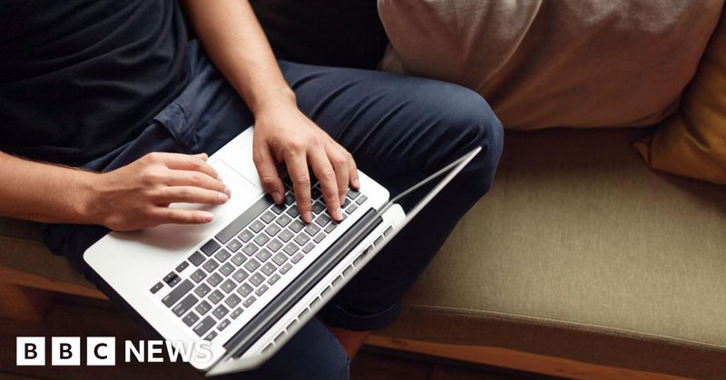 An unrecognisable man wearing a navy t-shirt and dark blue jeans is sitting on a sofa using a laptop.