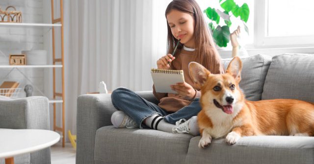 girl writing in a notebook, sitting on couch with her corgi