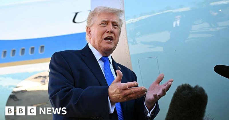Trump wearing a suit and blue tie gesturing with his hands in front of microphones