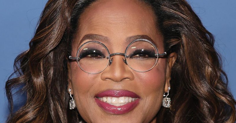 Oprah Winfrey smiling in round wire-rimmed glasses and a white jacket over a black V-neck shirt