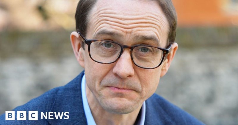 A close-up shot of BBC's political editor Chris Mason looking straight to the camera. He wears brown glasses, a blue shirt and blue blazer.