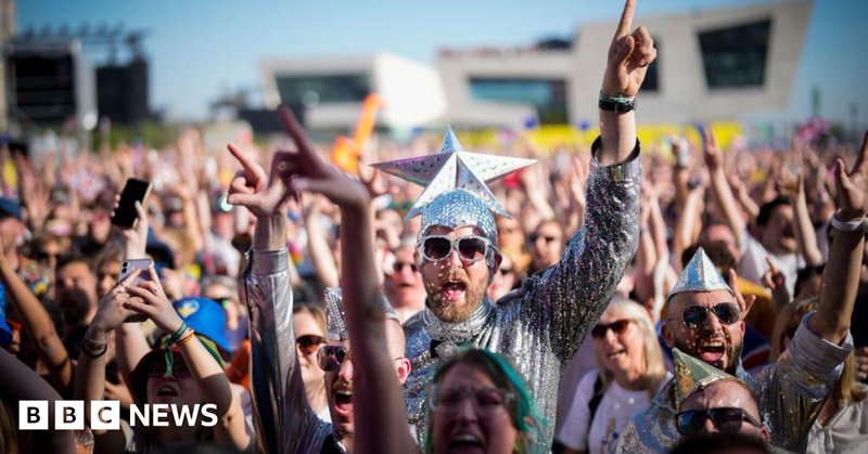 Eurovision fans watch the 2023 Eurovision Song Contest final on a giant screen in host city Liverpool. Many fans are dressed up, including the man in the centre of the photo who wears a silver jumpsuit and sunglasses and wears a silver cap with a giant silver star on the top.