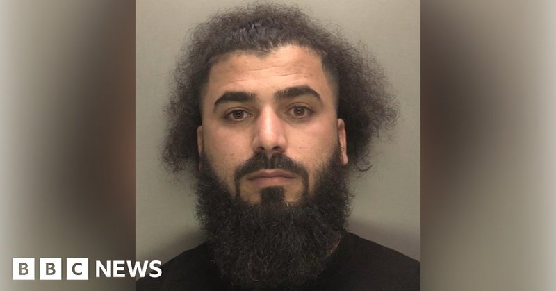 A mugshot of a man with a long fluffy beard and dark hair. He wears a black t-shirt and looks at the camera.