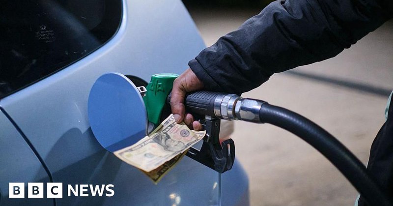 A hand holding a petrol pump into a car as it fills up at a petrol station in Zimbawe. The same hand also holds paper money. The car is silver.