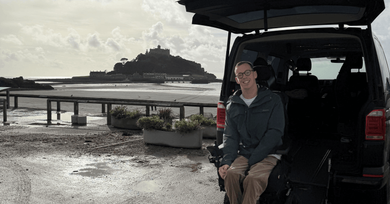 Keron Day sits in his wheelchair in front of the open boot of his Motability vehicle, with a sandy beach and an island in the background