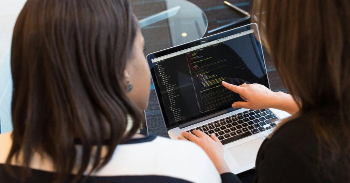 Two women look at software code on a laptop.