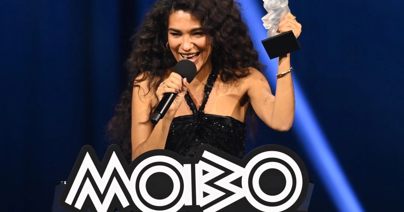 Olivia Dean, with long curly hair, smiles while holding a MOBO Award and speaking into a microphone.