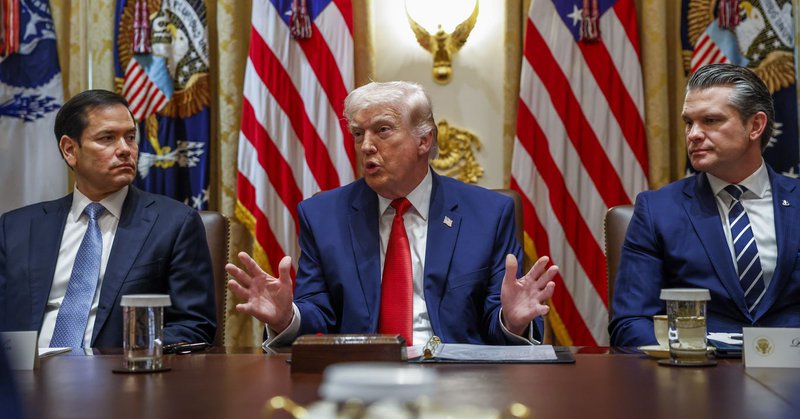 Three men in dark suits, Secretary of State, Marco Rubio (L), President Trump and Secretary of Defense Pete Hegseth, sit at a long wooden table in a formal room with multiple U.S. flags and gold decor; Trump, wearing a blue suit and red tie, speaks with his hands gesturing.