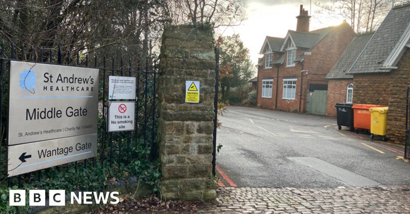 Entrance and gate lodge at a mental health hospital. In the foreground to the left are a number of signs. A cobbled road can be seen as well as multi-coloured bins. There are trees and a grey sky.