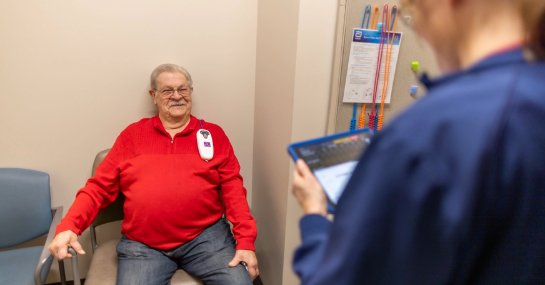 An older adult sits in a medical exam room chair while a healthcare worker stands nearby reviewing information on a tablet, with clinical equipment an