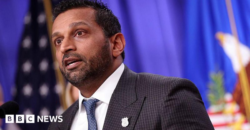 close-up of Kash Patel speaking, wearing a suit and tie in front of American flag