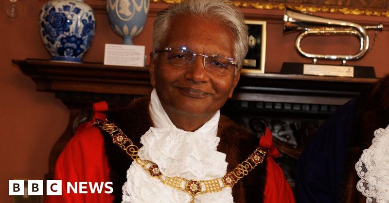 Wearing his mayoral robes and gold chain, Bharat Pankhania smiles at the camera. Behind him two vases and a musical instrument can be seen on a mantelpiece.