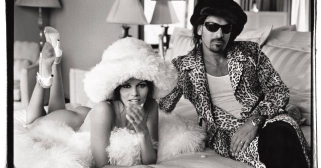 A black-and-white photograph shows a woman in a fluffy hat and heels lying on a bed beside a man in a leopard-print jacket and sunglasses in a hotel room setting.