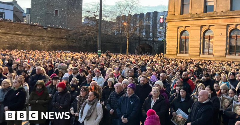 A large crowd of people stand side by side in Derry's Guildhall Square, a number are holding posters of a young woman.