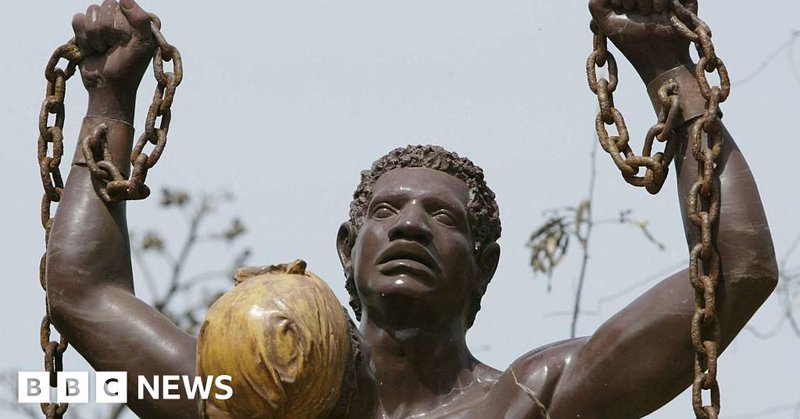 Close up of statue showing liberation from slavery on Goree Island in Senegal. It is a man from shoulders up, with his arms raised, holding a broken chain in his hands which is still attached to his wrists. The statue also includes the figure of a woman standing in front of the man - all that is visible of her is the back of her head, and she has a head wrap on.