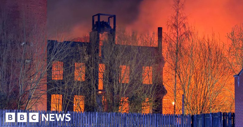 A large blaze at night in a mill building. The sky is lit up orange. Wooden fencing is in front of the building.
