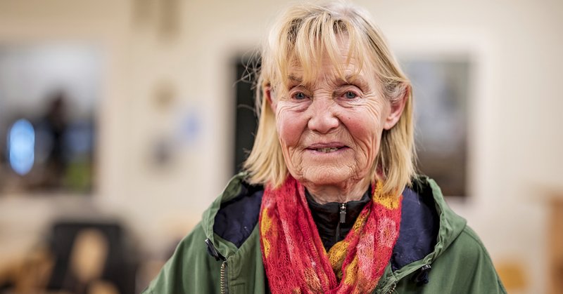 En äldre dam iklädd en grön jacka med en röd scarf runt halsen som tittar in i kameran.