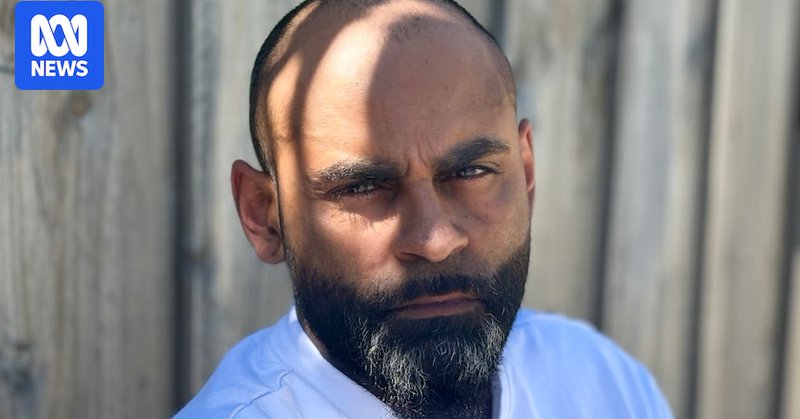 An Indigenous man in front of a wooden fence with mottled light over his face.
