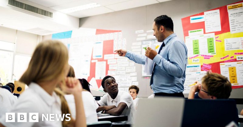 A stock image shows a secondary teacher standing teaching in a classroom wearing a suit and tie. The students are looking at him from their desks, wearing white short sleeved shirts.