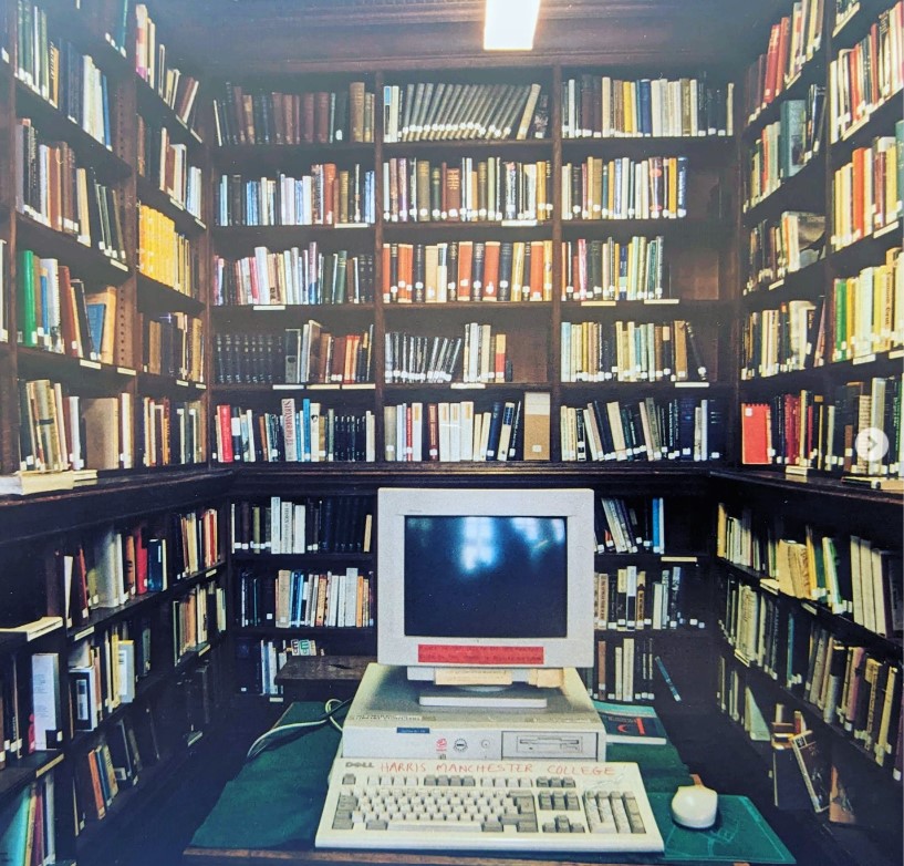 A room with books in bookcases on every side. In the middle is a desk with an old style computer on it (the screen is small, and the hard drive is in a box under the screen).