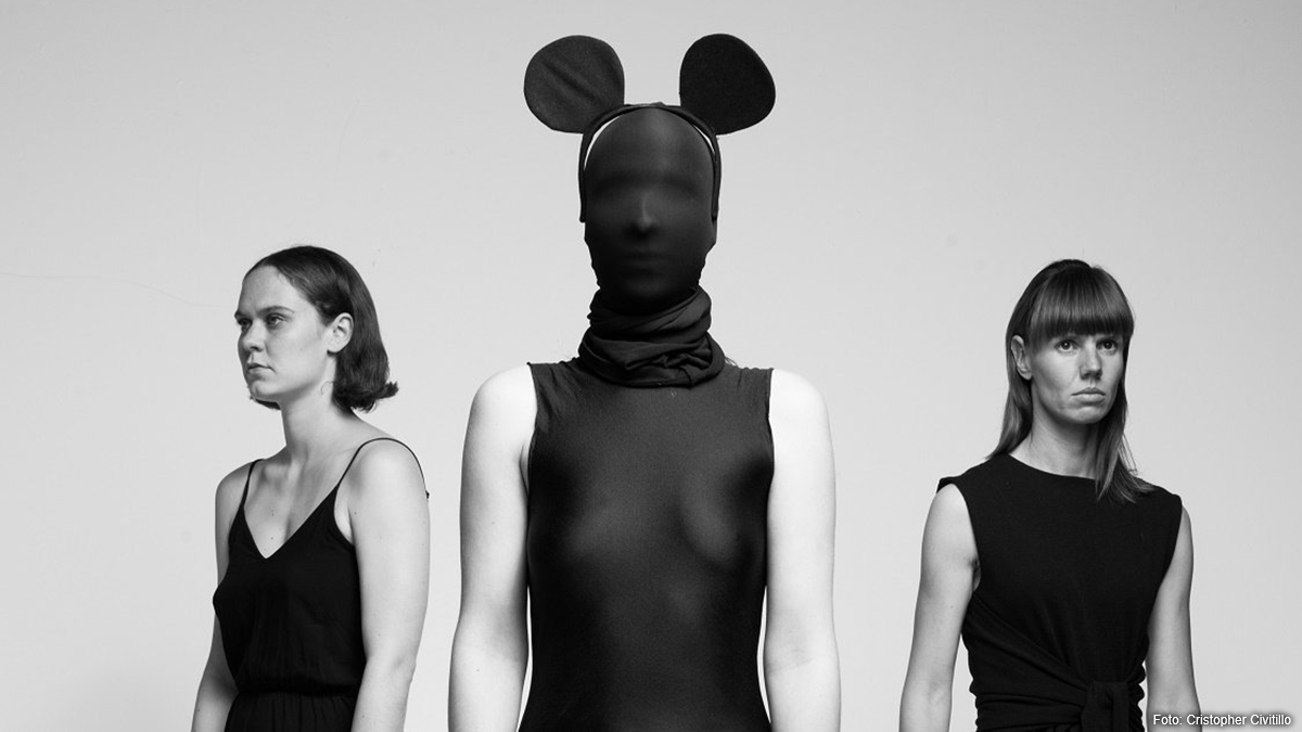 Mittig eine Person mit schwarzer Maske, die das ganze Gesicht bedeckt und Mickey Mouse Ohren hat, links und rechts die Tänzerinnen Eva Borrmann und Evelyn Hornberg, die zur Seite schauen