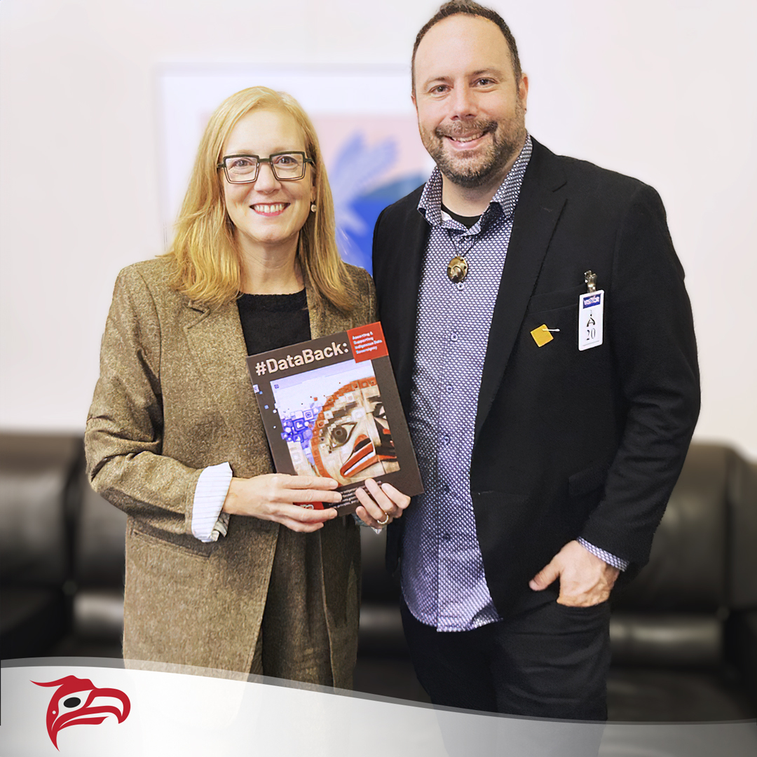 Photo of Jeff Ward, CEO of Animikii, standing to the right of Brenda Bailey, BC's Minister of Jobs, Economic Development and Innovation. They are indoors, smiling at the camera, holding a copy of the #DataBack eBook, with the Animikii logo in the background. Jeff is wearing a dark blazer and Brenda is in a tweed blazer, promoting the importance of Indigenous Data Sovereignty.