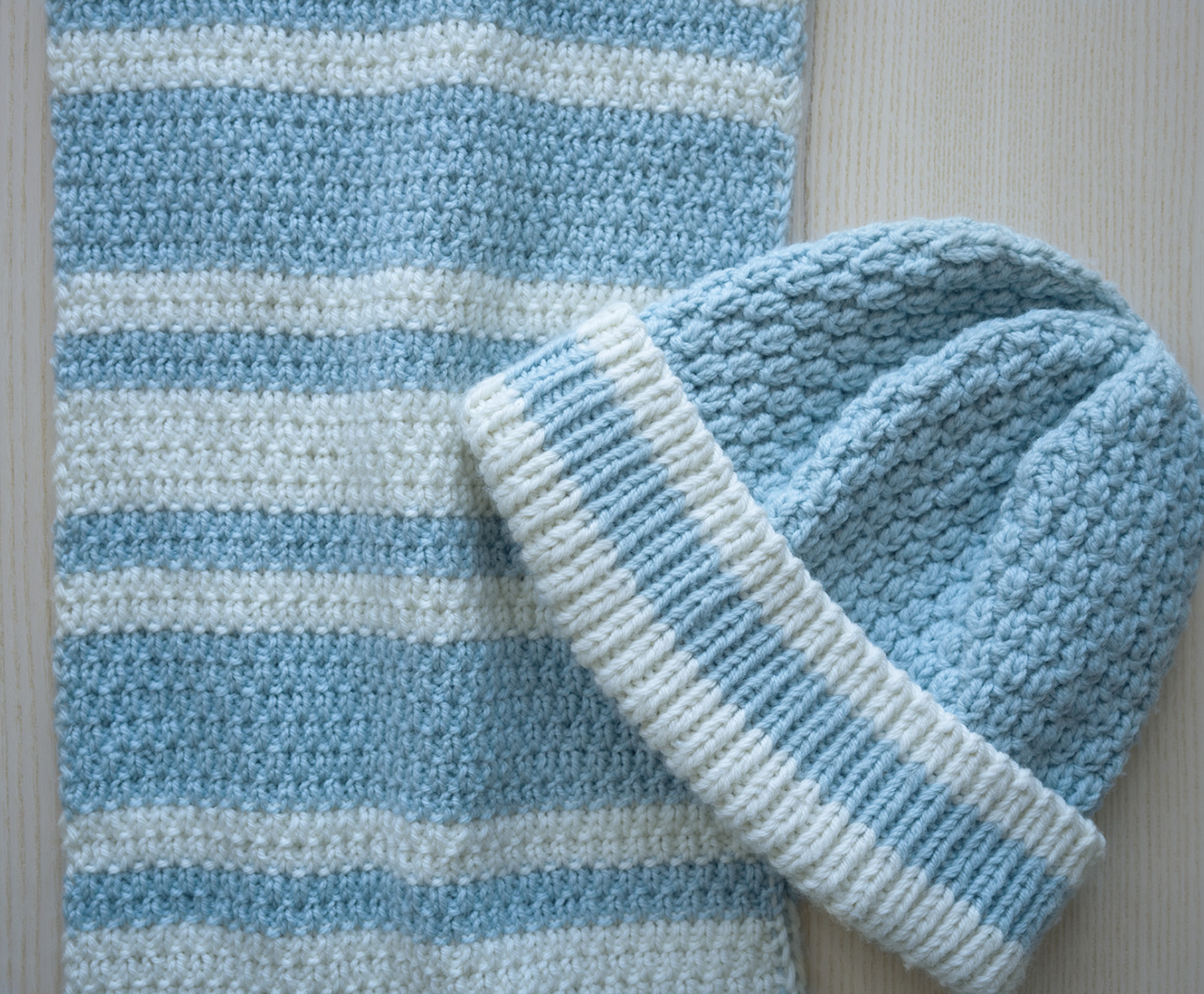 The photo shows a scarf worked in a light blue wool yarn with contrast white stripes and a co-ordinating hat. The scarf is a light-weight version of the Petersfield Scarf by Moira Ravenscroft, Wyndlestraw Designs.  