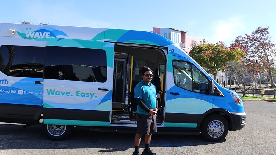 A blue and green MTD "The Wave" van parked in a lot with the side door open, showing the interior, while a driver stands beside the van.