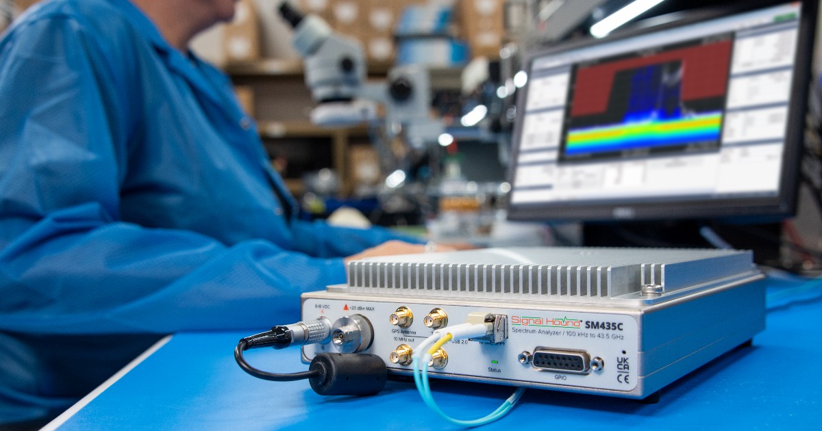 A person working in a laboratory, using a Signal Hound SM435C spectrum analyzer connected to a laptop displaying colorful spectrum analysis graphs.