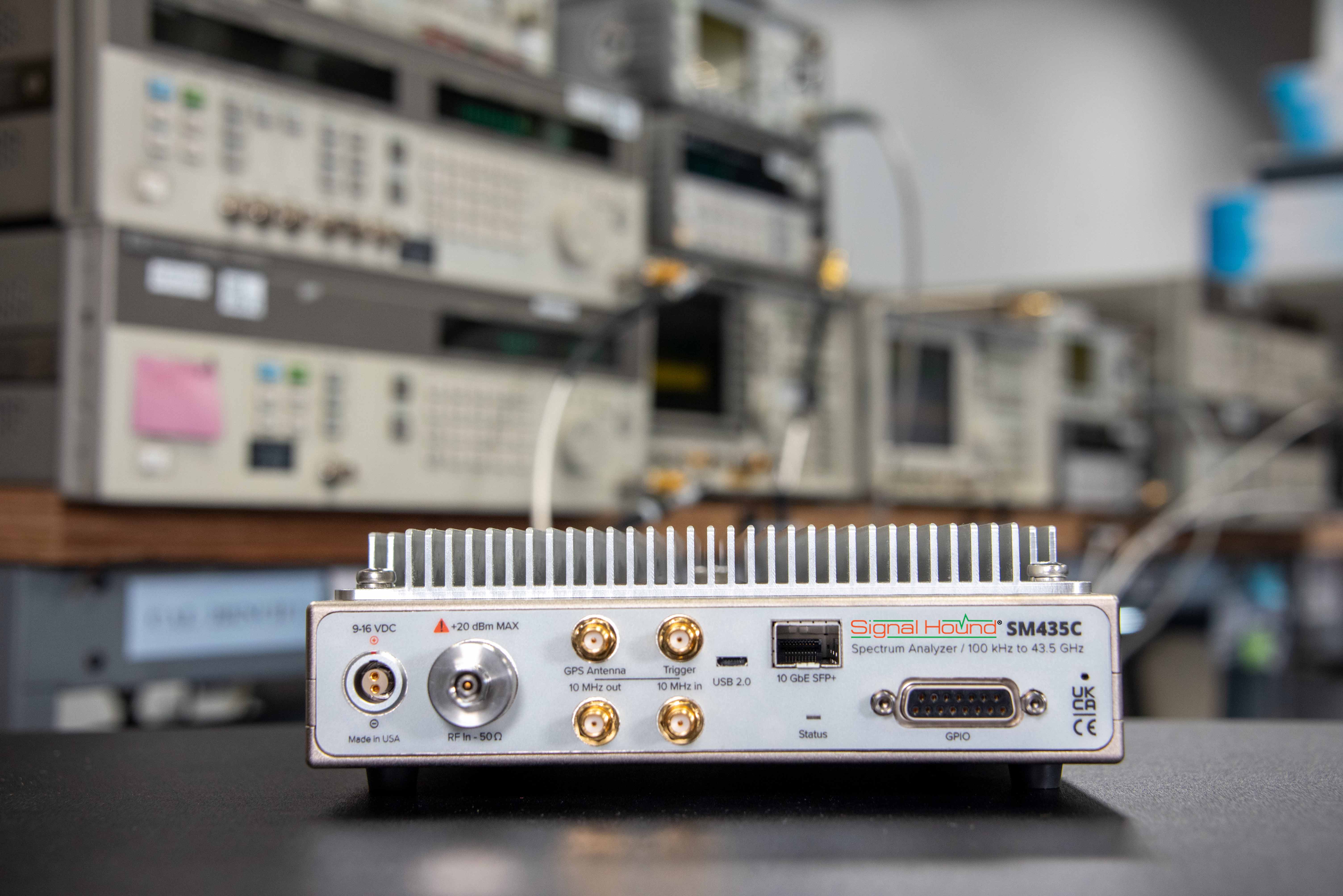 A Signal Hound SM435C spectrum analyzer device in focus, with various pieces of electronic testing equipment blurred in the background.