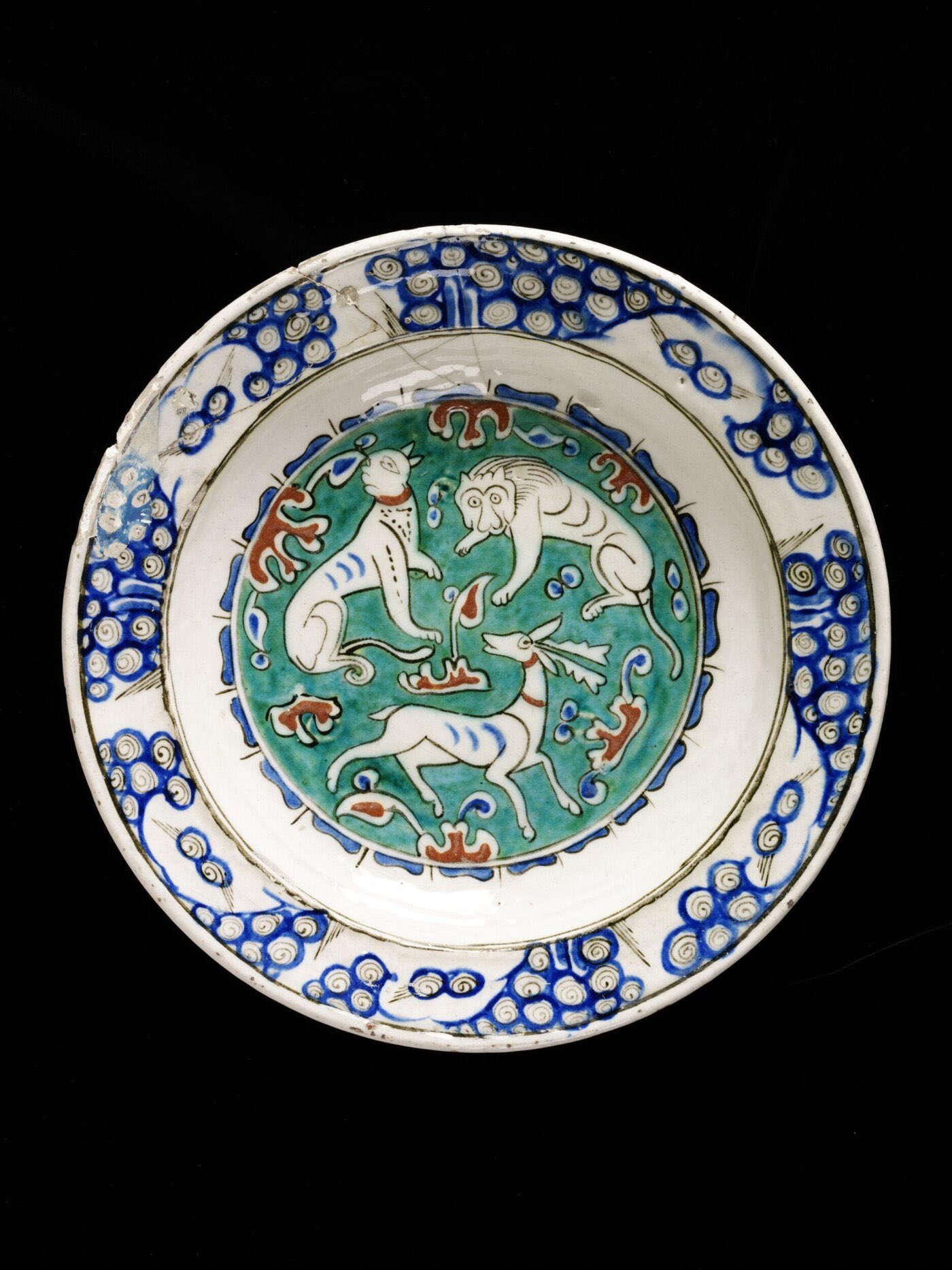 “Dish of glazed greyish fritware, and painted with a green medallion containing a lion, dog and stag in white, and the intervening spaces being filled in with red and blue ornament devices. The rim is decorated with groups of olive-green spirals on a blue ground.”
“Fritware, polychrome underglaze painted, glazed”