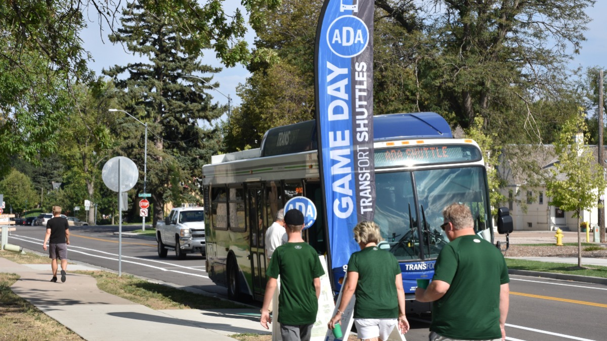 A bus parked on a street with a flag beside it that says GAME DAY TRANSFORT SHUTTLES. Three people are walking toward the bus.