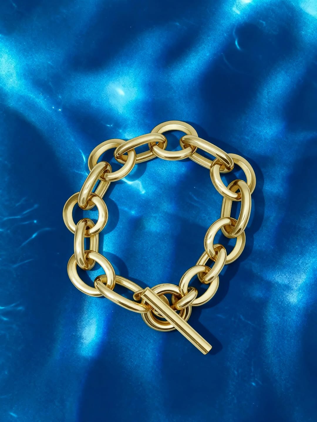 Shiny gold toggle bracelet with oval links and a toggle clasp resting on a rippling blue water surface.