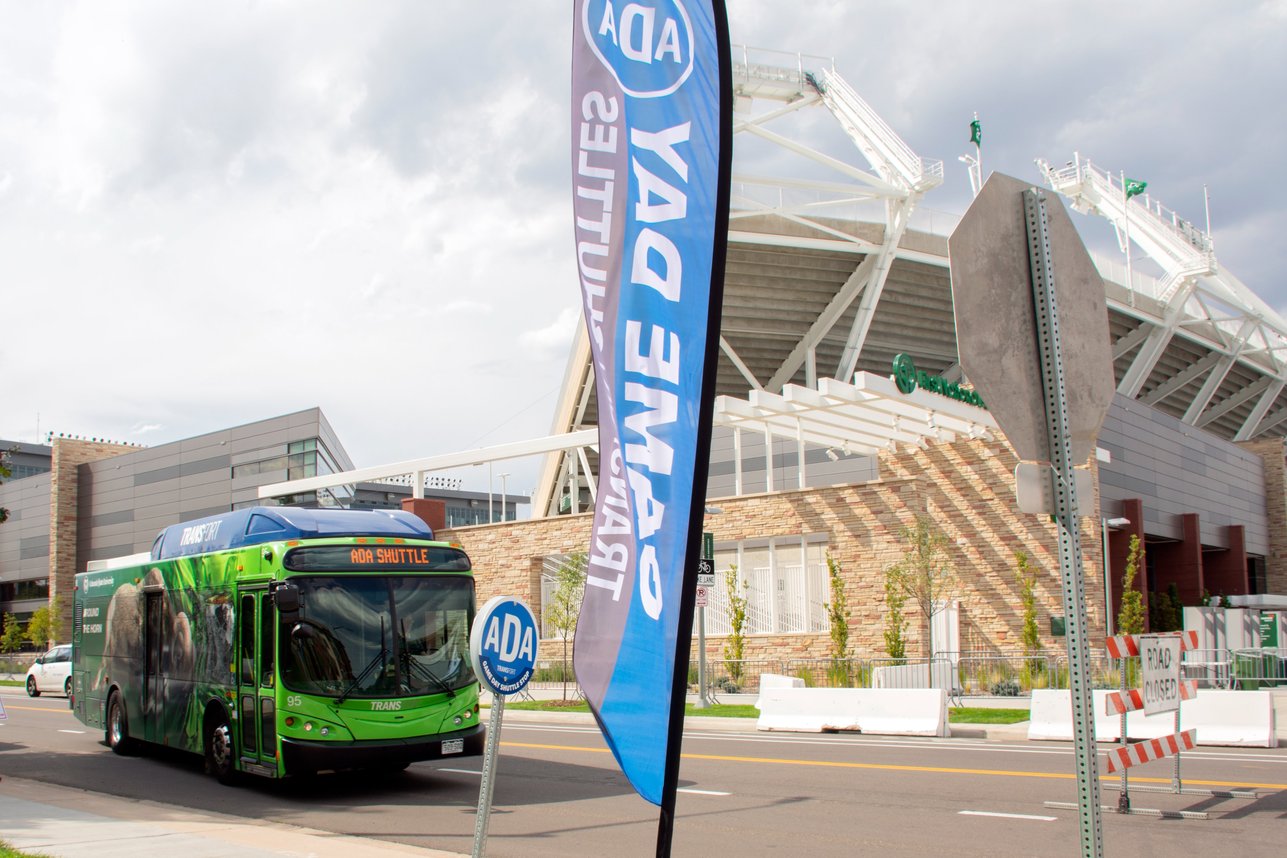 A green bus driving past a stadium with a flag flying by the sidewalk that says Game Day Shuttles.