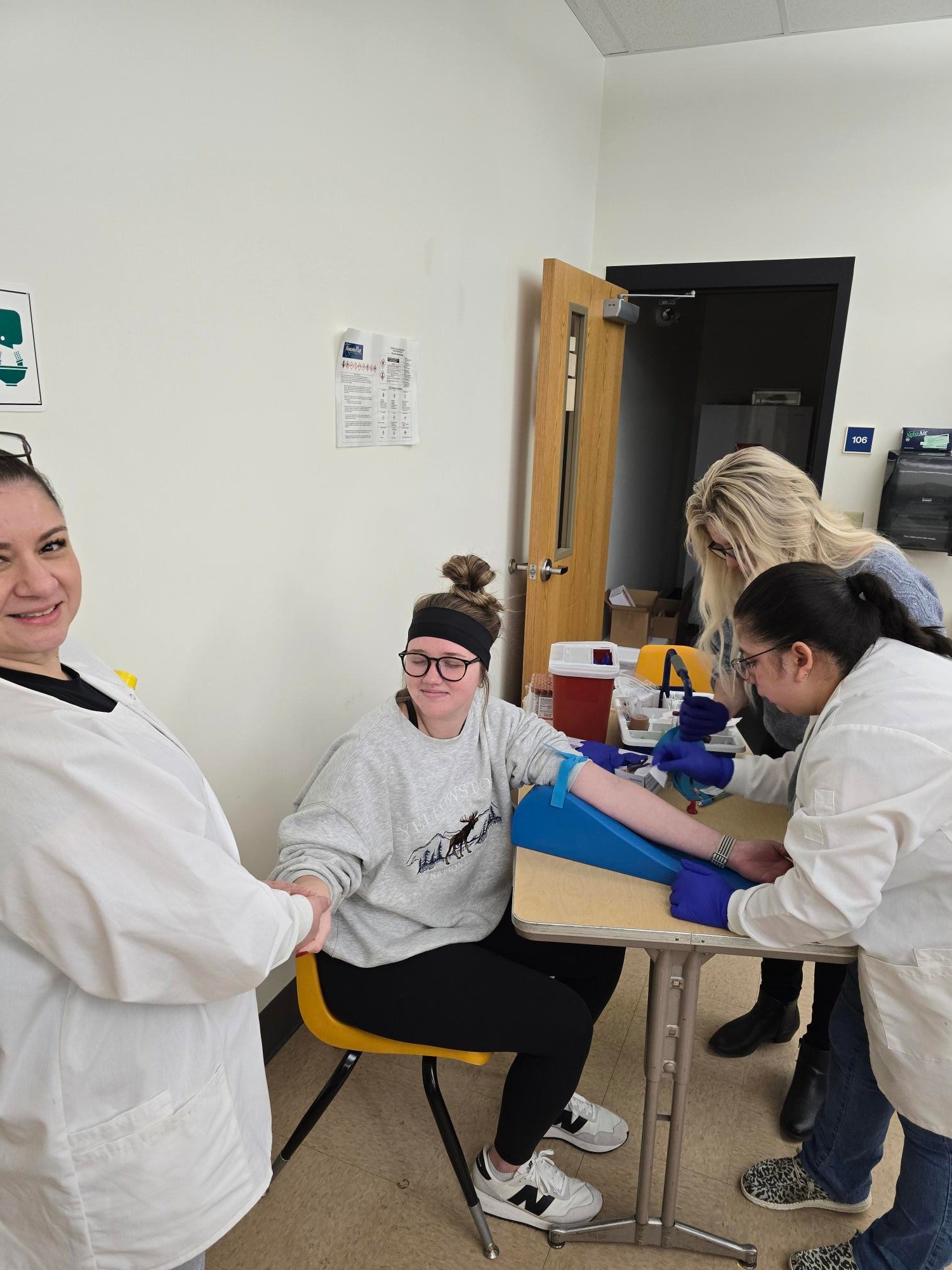 A person seated at a table with an arm extended while two individuals in lab coats prepare for a blood draw in a clinical setting. Another person stands nearby observing.