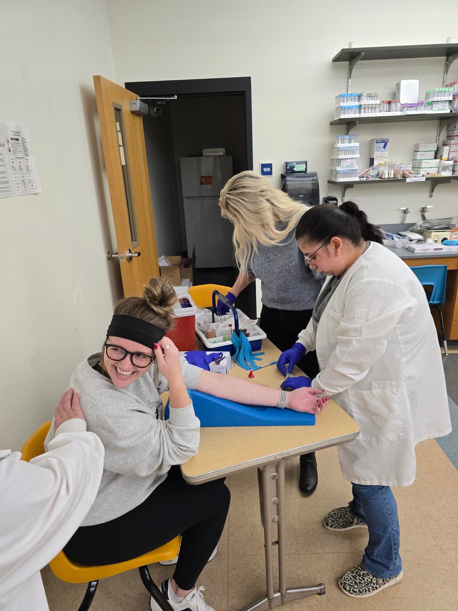A person seated at a table during a blood collection procedure, with two individuals in lab coats assisting and medical supplies visible on the table.