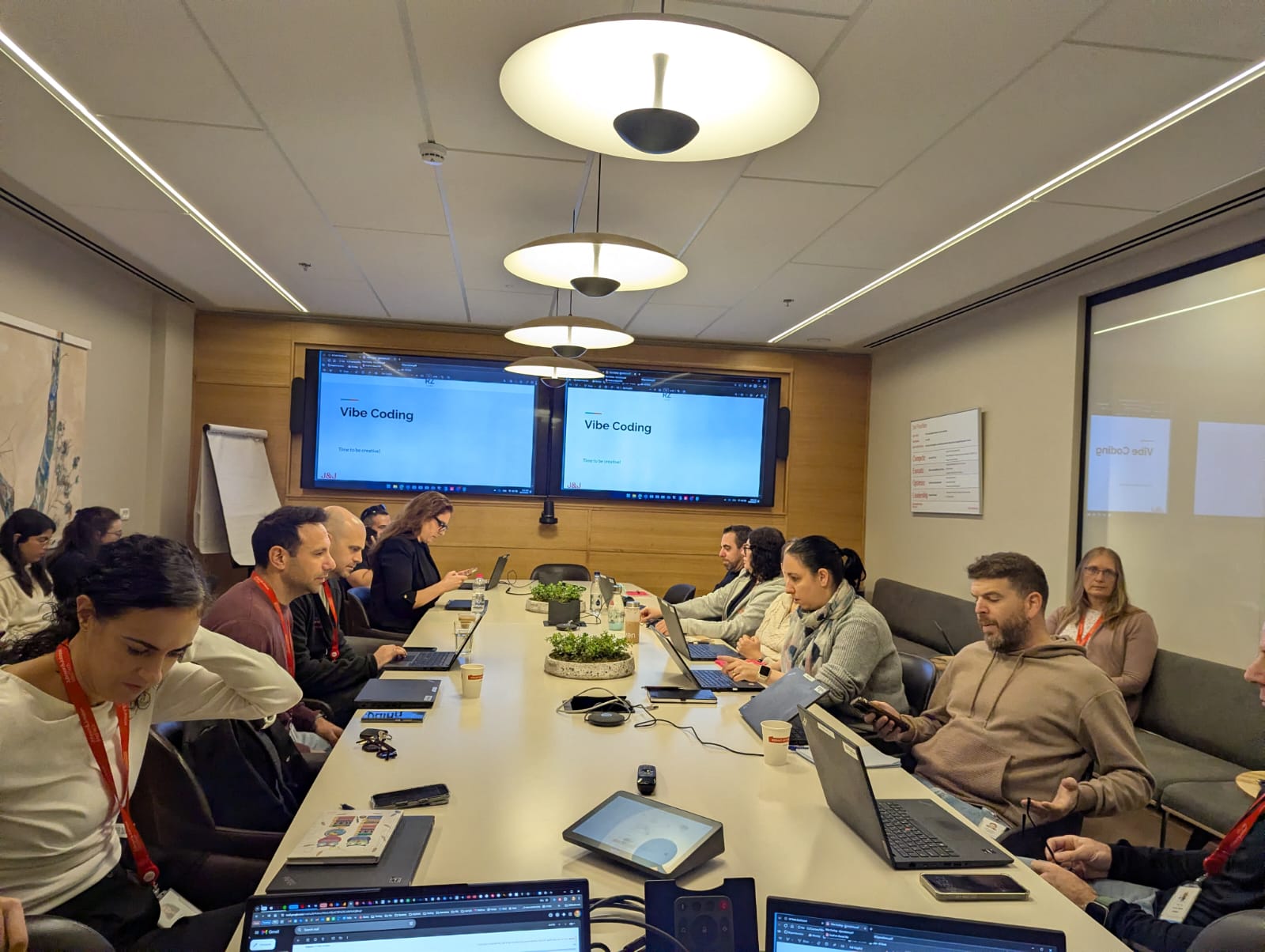 A panoramic shot of the Vibe Coding workshop in session. Ten participants from the Johnson & Johnson MedTech Operations team are seated around a long, well-lit conference table, engaged in coding exercises on their laptops. Two large, mounted screens at the far end of the room prominently display the title 'Vibe Coding'.