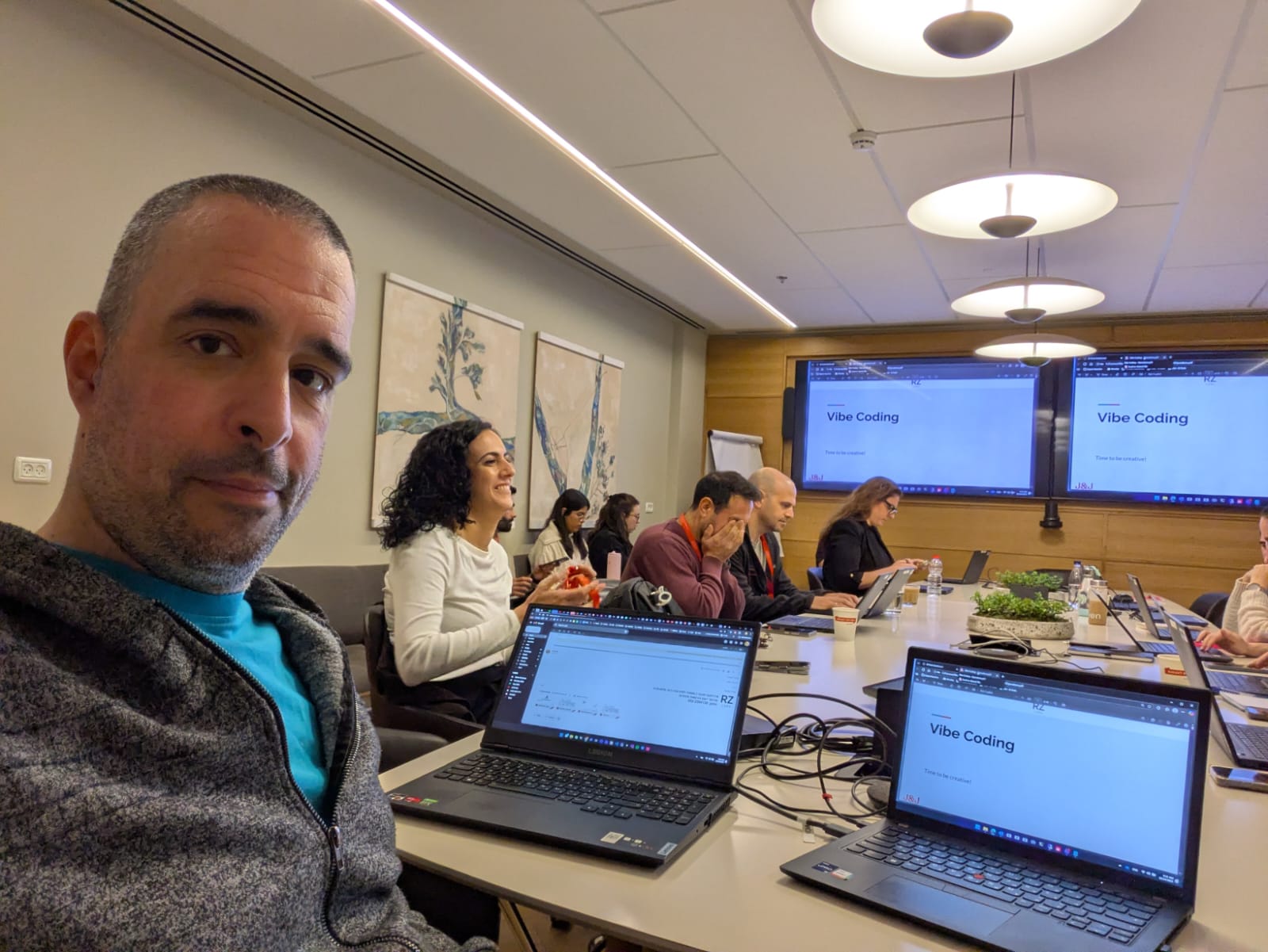 A wide selfie photo taken during a Vibe Coding workshop at Johnson & Johnson MedTech. The instructor is in the foreground looking at the camera. A group of approximately ten participants are seated around a long conference table working on their laptops. The title 'Vibe Coding' is visible on multiple external monitors in the background.