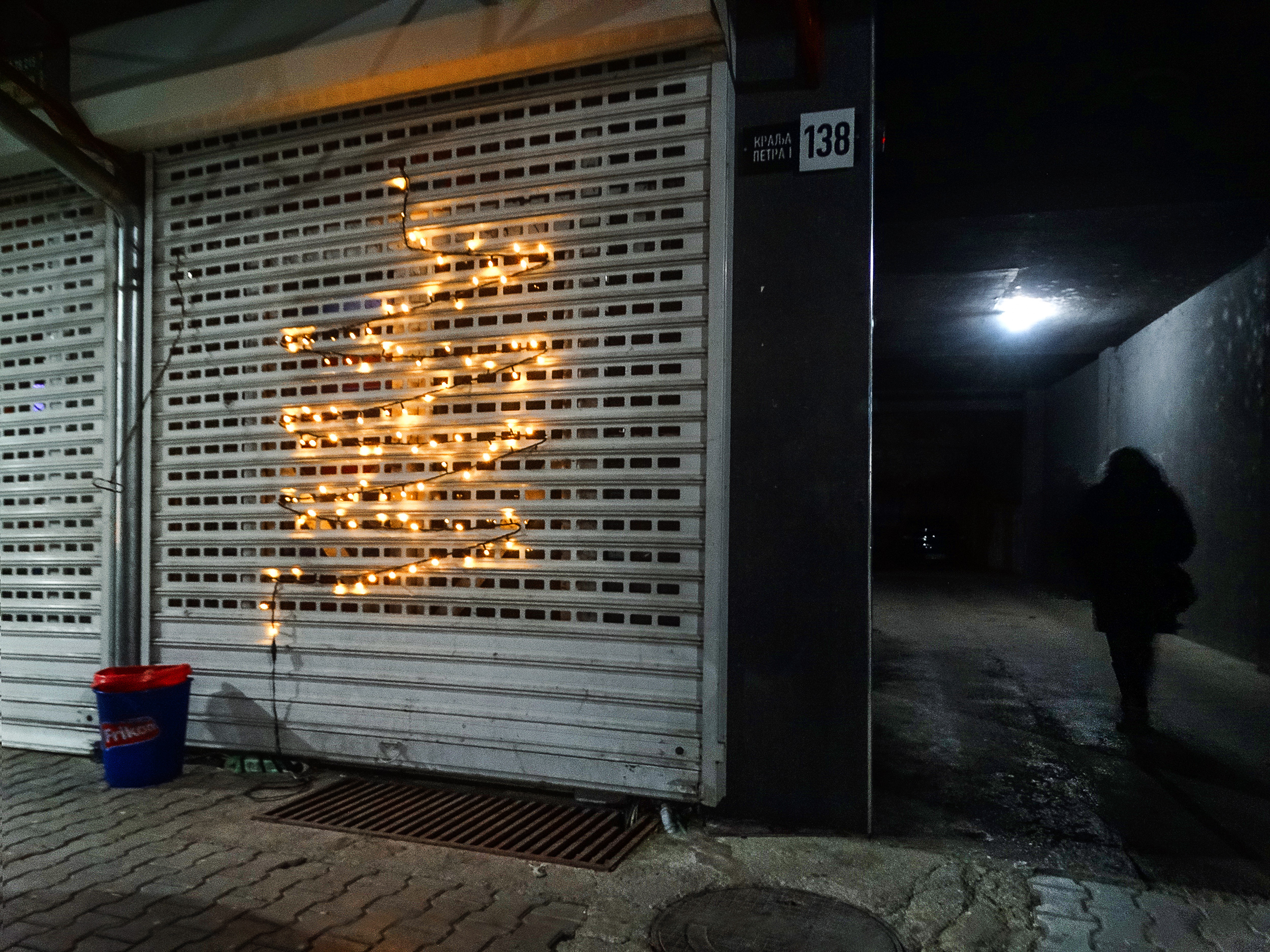 Night-time black and white street documentary photograph taken on December 31, 2025, in Zvečan, Kosovo and Metohija. An improvised Christmas tree made of string lights is attached to a closed shop shutter, while a solitary human silhouette walks through a dark passageway. Photograph by Ivan Maksimović, Serbian documentary and street photographer from Kosovo and Metohija.