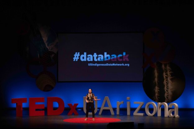 Stephanie Russo Carroll stands on a stage with large block letters behind here reading "TEDx UArizona". A screen above her displays a graphic reading "#databack".