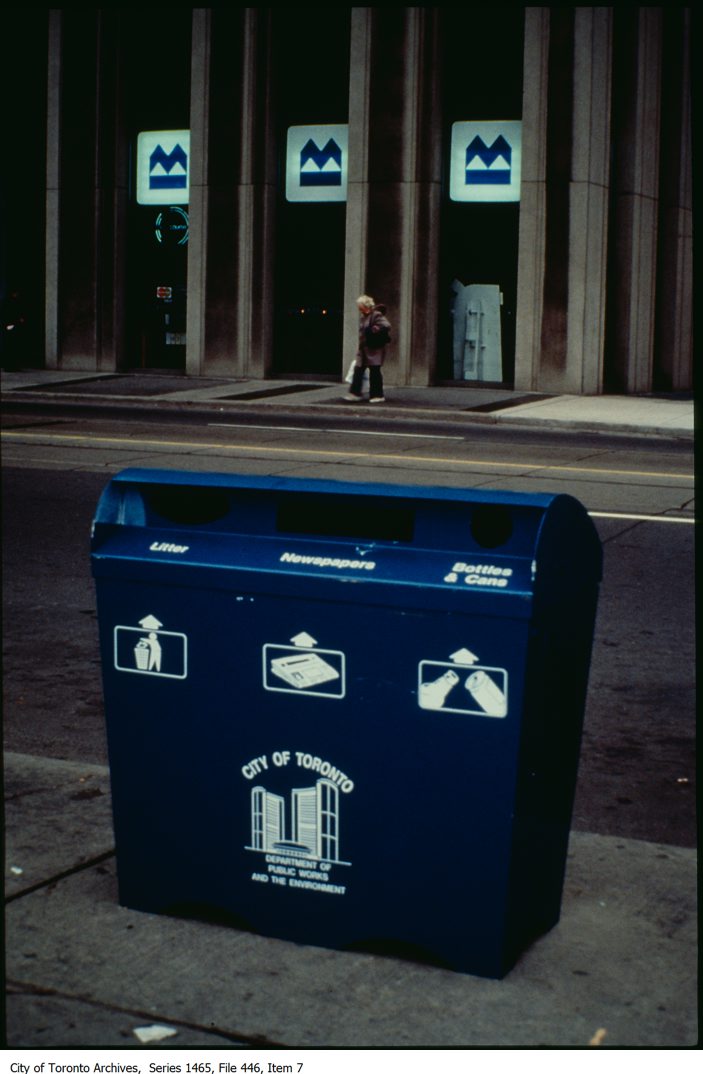 A colour photograph of a blue garbage/recycling bin on the side of the road. There are three inserts, one for litter, one for newspaper, and one for bottles and cans. There is an image representation for the three categories and below that is a City of Toronto logo. Across the street is a building with three Bank of Montreal logos on display. Series 1465, file 446, item 7.