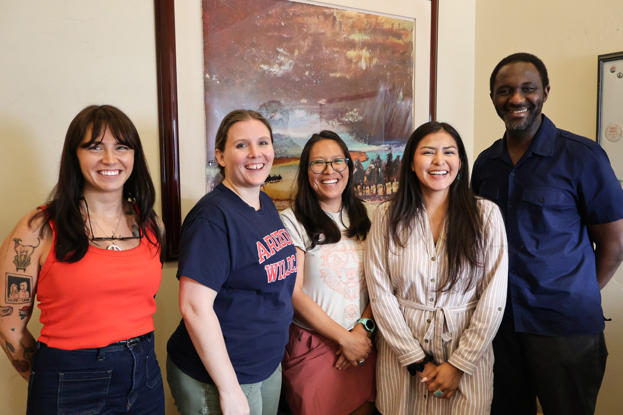 Collab members pose in the Udall Center lobby in front of a large abstract landscape painting. Pictured from left to right are:
- Cassidy Schoenfelder (Oglála Lakȟóta), PhD Candidate in the School of Geography, Development, and Environment (SGDE); Collab Research Associate & SGDE Graduate Associate
- Jewel Cummins, PhD Candidate in American Indian Studies; NNI and Collab Graduate Research Assistant
- Bree Lameman (Diné), PhD Candidate in the Mel and Enid Zuckerman College of Health (MEZCOPH); Collab Graduate Research Associate
- Caleigh Curley (Diné, Hopi), PhD Candidate in the Public Health Policy and Management program at MEZCOPH; Research Program Administrator at the MEZCOPH Community, Environment, and Policy department
- Ibrahim Garba (Karai-Karai), Assistant Research Professor at MEZCOPH, NNI Senior Researcher